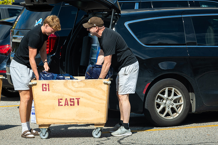 A older man helps a younger man load a wooden cart from the back of a car
