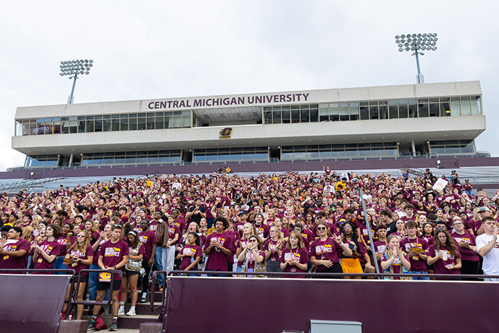 A large group of young people stand in the seating of a football stadium with the words Central Michigan University written across the top of the clubhouse