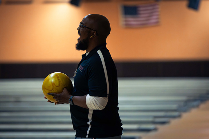 A man with a beard and glasses laughs while holding a bowling ball with bowling alleys in the background