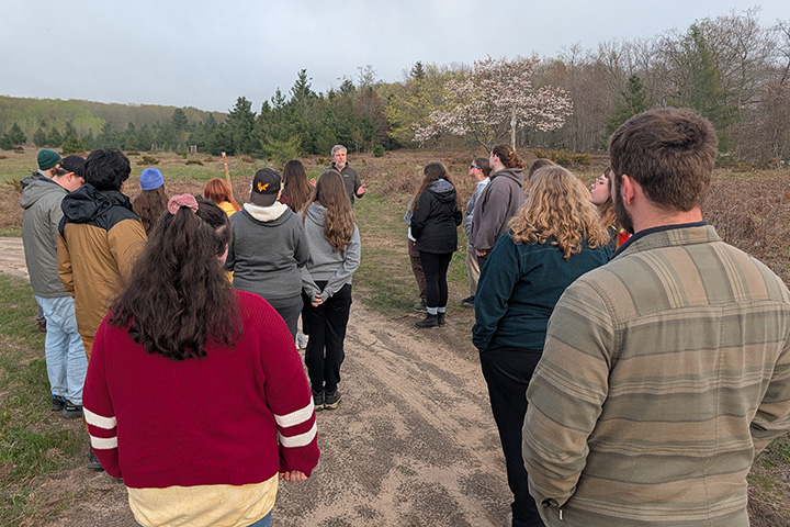 Several young people wearing jackets and flannel shirt stand around a bearded man who is lecturing to them