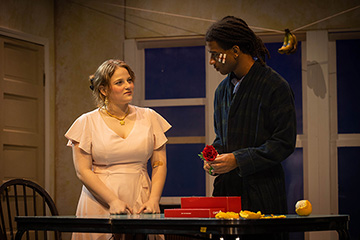 A young woman wearing a peach colored dress with her hair tied in buns speaks with a young man in dark clothing with two bandages on the side of his face in front of indoor play scenery