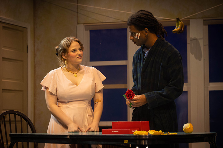 A young woman wearing a peach colored dress with her hair tied in buns speaks with a young man in dark clothing with two bandages on the side of his face in front of indoor play scenery