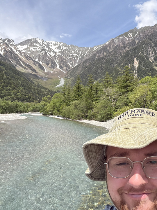 A closeup selfie of a male college student wearing a hat with a river and mountain in the background