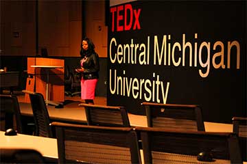 A Black woman in a pink skirt stands in front of a large screen that reads TEDxCentral Michigan University