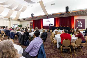 A room full of people sitting at round tables listens attentively to a man speaking onstage in front of a white screen that says Go Grants