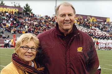 A woman in a yellow sweater and a man in a maroon coat stand on a football field