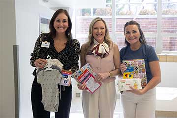 Three women smile into the camera while holding donated baby care items