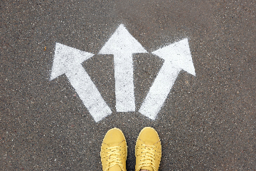 A top-down view of two yellow shoes standing on pavement. Three white arrows painted on the pavement near the tips of the shoes point in different directions.