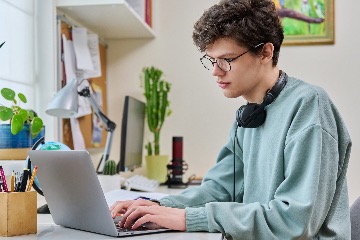 A male high school-aged student wearing glasses and headphones sits at a laptop computer while at a desk in a well-lit bedroom.