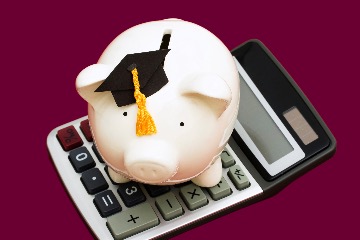 A piggy bank with a graduation cap sits on top of a calculator in front of a maroon background.