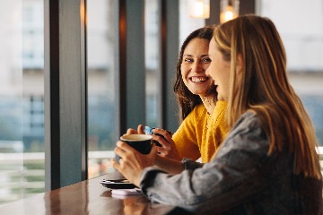 Two young women sit at a table drinking coffee and smiling. The room is surrounded by large windows with bright, natural light brightening the room.