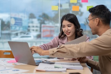 A man and woman sit at a table covered in papers and notebooks pointing at a laptop and talking together.