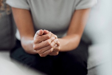 A closeup of a woman from the shoulders down. She sits on a chair, hands clenched and tense.