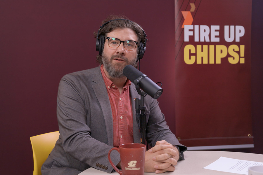 A bearded man wearing glasses, a sport coat, red shirt and headphones sits in front of a microphone at a table. A Fire Up Chips banner hangs on a maroon backdrop in the background.