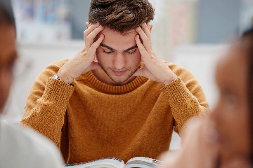 A young man in a mustard yellow sweater holds his hands to his temples looking down towards a book with his eyes closed.