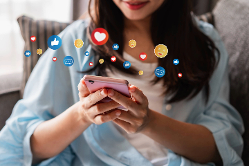 A young woman sits in a chair using a mobile phone in a pink case as superimposed social media icons float around.