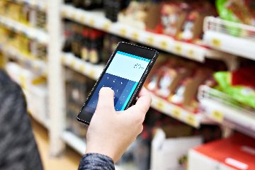 Inside a supermarket, a closeup of a person's hand holding a phone with a calculator screen on display. Food products on shelves are blurred in the background.