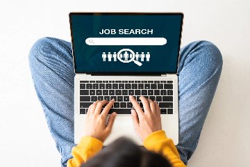 A top-down view of a person wearing jeans and a yellow long sleeve shirt shows the person typing on a laptop with the words "Job Search" on the screen.