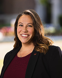 Female with long brown hair wearing a black blazer and maroon shirt smiles outside for her headshot.