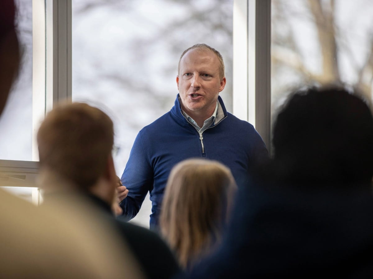A man in a quarter zip speaking to a group of people indoors.