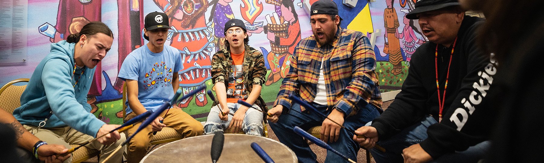 5 men singing and hitting a drum with sticks in front of a colorful background