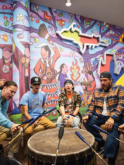 5 men singing and hitting a drum with sticks in front of a colorful background