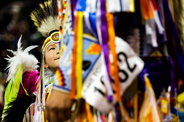 A young Native American boy in traditional regalia looks at his parent who is also in multicolored regalia.