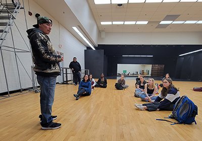 Man in jeans and a sweatshirt standing in a room with a wood floor where students are sitting.