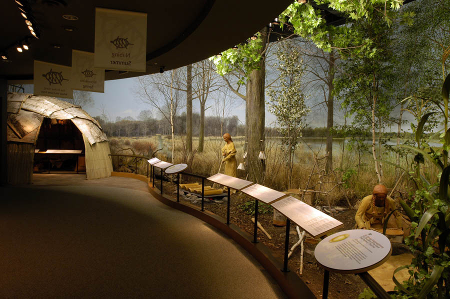 Exhibit within the center a lodge made from birch bark and reads on the left. A woman in buckskin making maple syrup in the middle and a man in buckskin on the right bent over with a pan of wild rice.