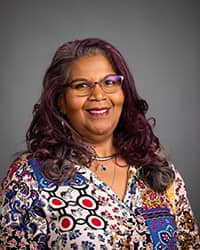 Elizabeth Husbands, CSID Dedicated Counselor profile picture. They are wearing glasses, hair down with curls, multi-colored blouse and a grey background