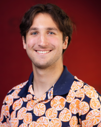 A young man smiling in front of a maroon background, wearing a shirt a blue collar and printed oranges