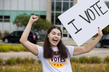 Picture of student smiling and holding up a vote sign