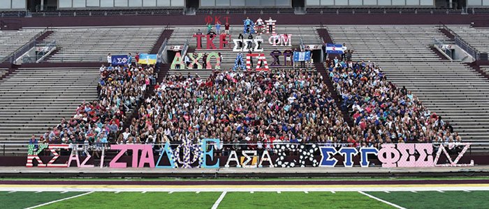 CMU fraternities and sororities pose with greek letters while sitting in stands at football stadium