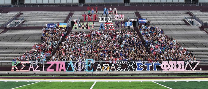 CMU fraternities and sororities pose with greek letters while sitting in stands at football stadium