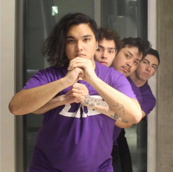 Four men with dark hair and wearing purple t-shirts stand in a line front-to-back with arms interlocked