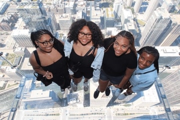 A group of women posing for a picture while standing in a sky scraper that has a clear floor and there are buildings underneath them.