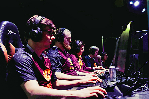 Students wearing headsets and maroon and gold uniforms sit at gaming computers