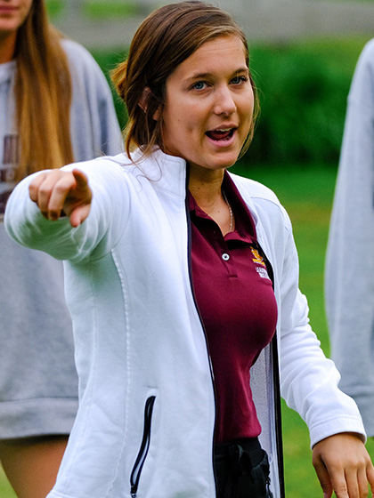 Student wearing a white jacket and maroon shirt, providing instruction to others outdoors