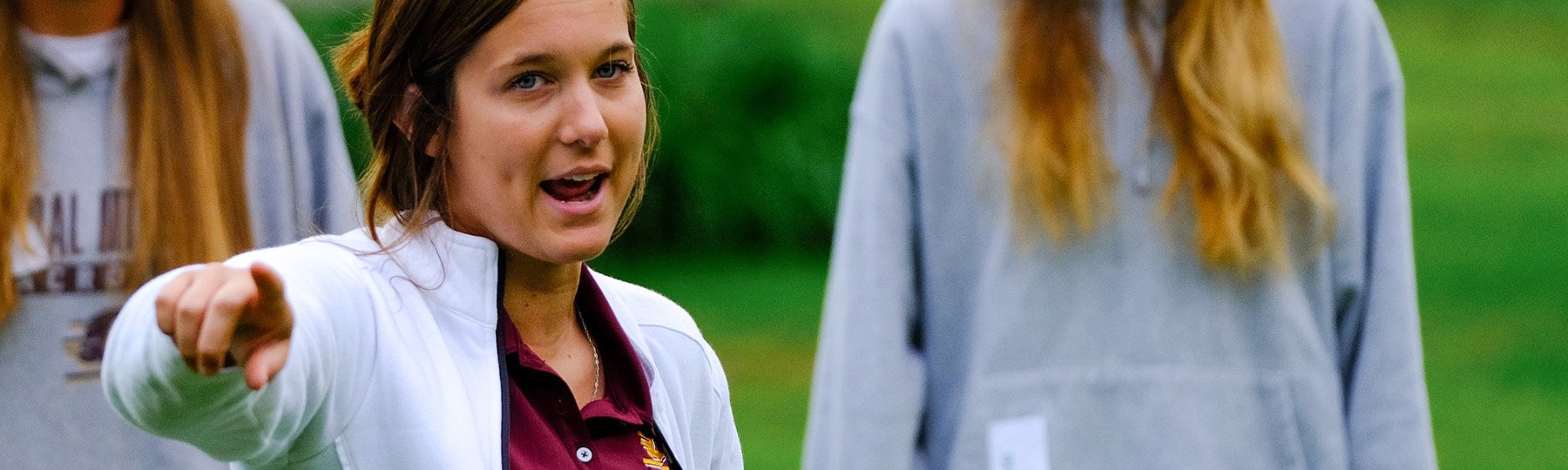 Student wearing a white jacket and maroon shirt, providing instruction to others in an outdoor setting