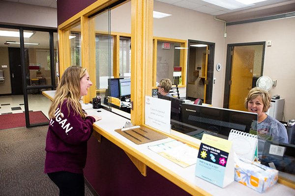 A student in maroon shirt interacting with a woman at a desk and computer.