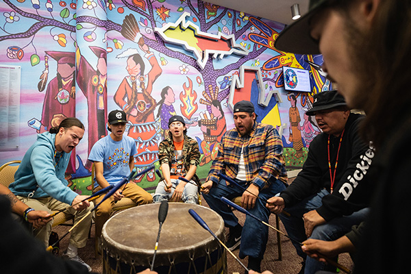 Indigenous people singing and playing the drum in front of a colorful mural