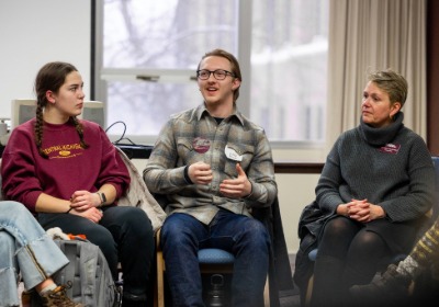 A group of people sitting in chairs with a student wearing glasses speaking.