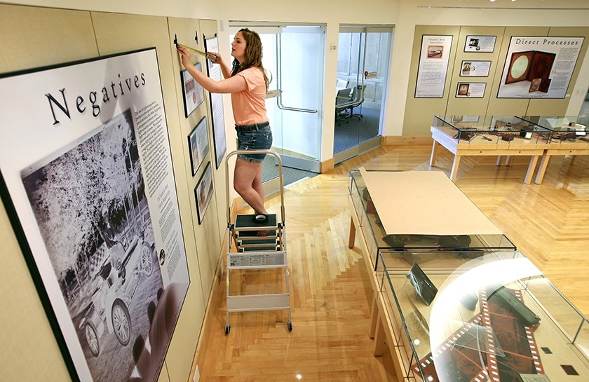 A person standing on a ladder to install photography exhibit in the Clarke Historical Library