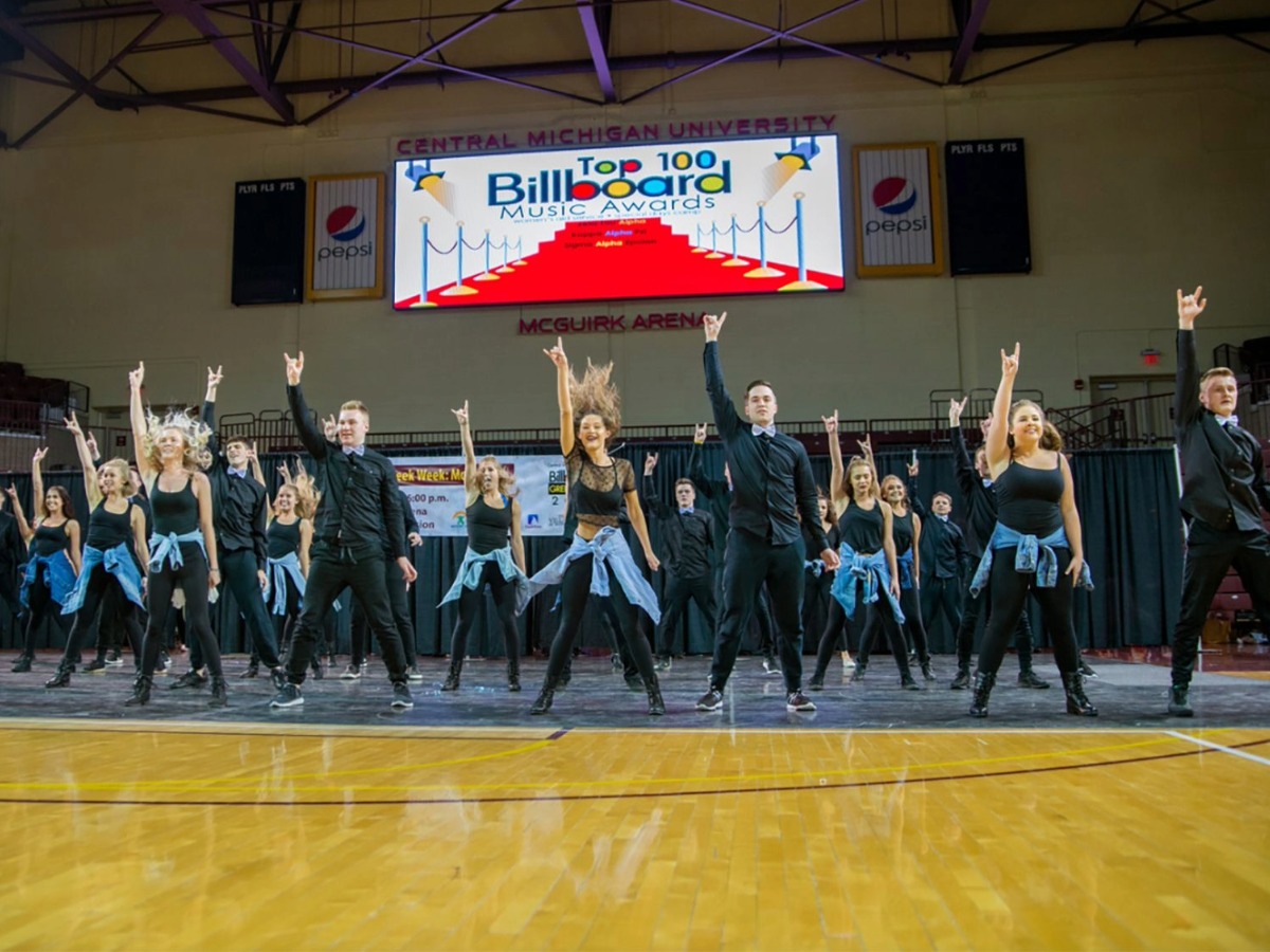 a large group of performers poses during a dance routine, with an arm raised and a hand making the rock symbol