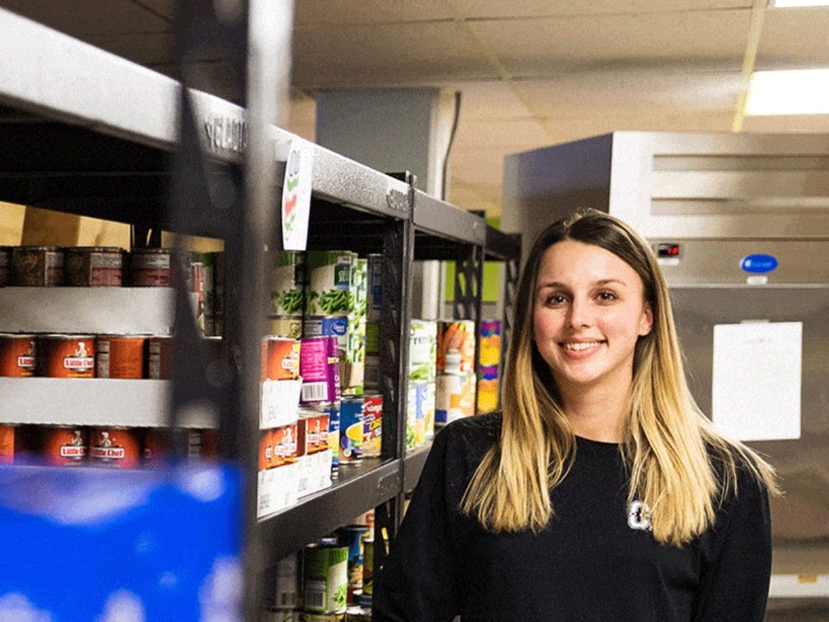 student volunteer smiles next to a stocked shelf of canned goods