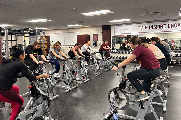 A cycling class with about a dozen people on stationary bikes in the MP-Fit room at the Student Activity Center