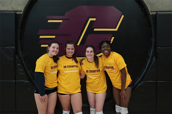 Four female intramural volleyball players in bright yellow "IM Champion" tee shirts stand in front of a giant Central Michigan "action C" logo.