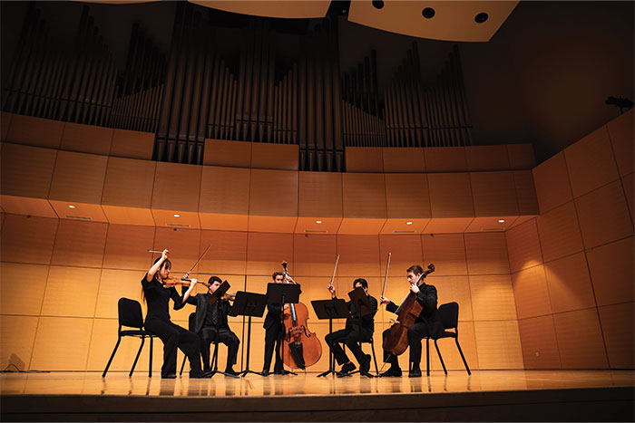 2018-486-10-Music-Feature-2-as_700x467 A quintet of five string players sit on stage, performing. Several of the players have their bows raised in the air, mid-note.