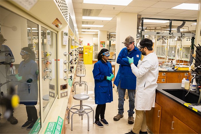 2022-181-0162-BioChemistry Labs 1 700x467 Three students stand in a chemistry laboratory talking. All three students are wearing lab coats, safety goggles and gloves.