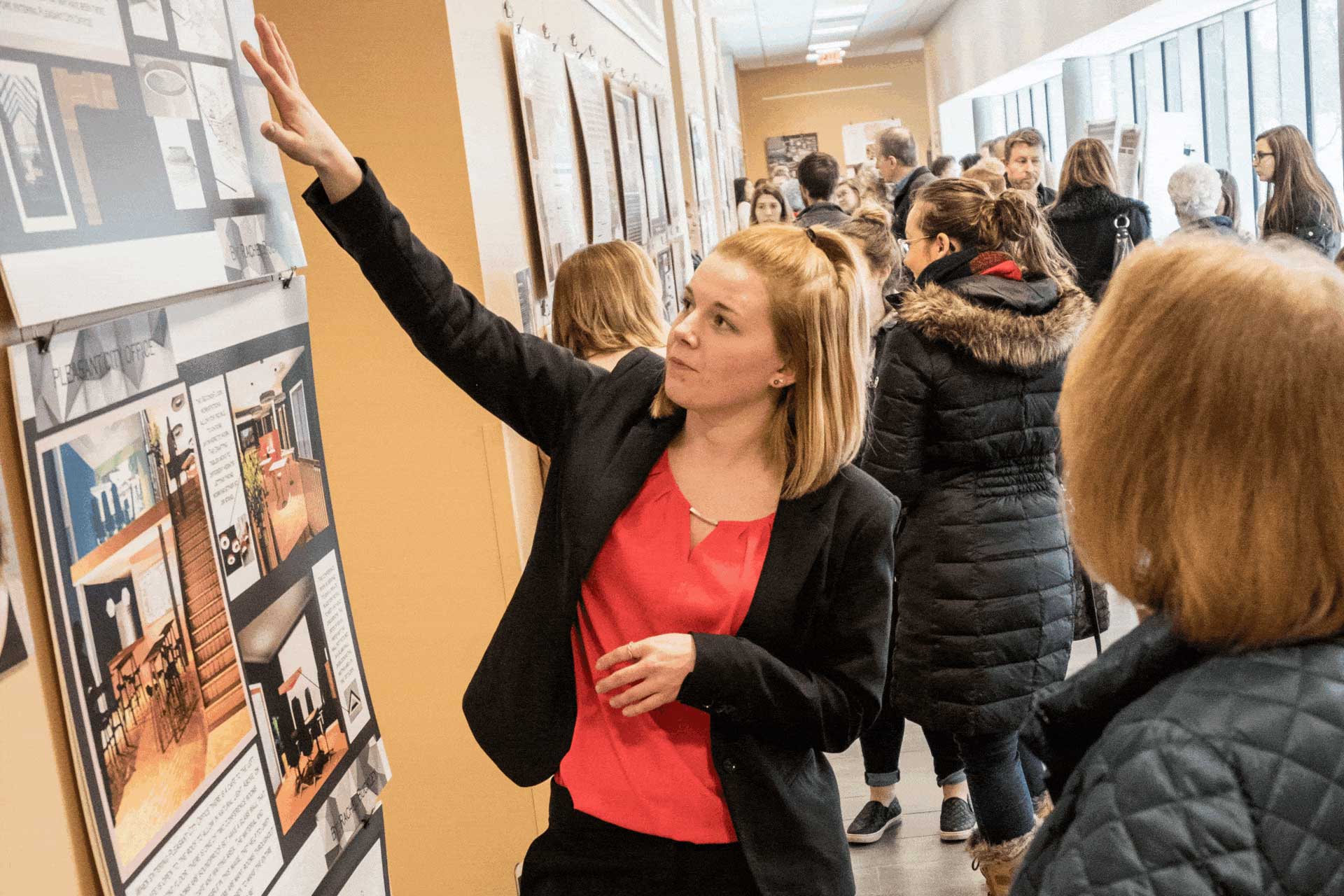 Lot of people walking in the hallway and students explaining the various interior design which is hung up on wall